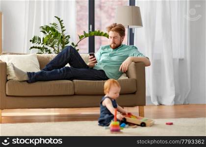 family, fatherhood and technology concept - red haired father with smartphone and little baby daughter playing with toy blocks kit at home. father with smartphone and baby playing at home