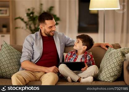 family, fatherhood, technology and people concept - happy father and little son with tablet pc computer sitting on sofa at home in evening. father and son with tablet pc playing at home