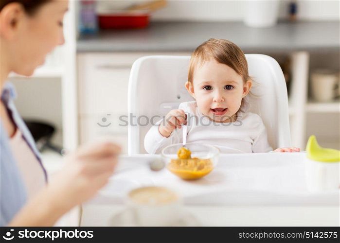 family, food, breakfast and people concept - happy mother with coffee cup and little baby sitting in highchair and eating puree at home kitchen. happy mother and baby having breakfast at home
