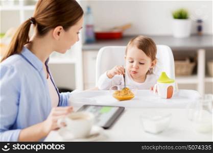 family, food, breakfast and people concept - happy mother with tablet pc and little baby sitting in highchair and eating puree at home kitchen. happy mother and baby having breakfast at home