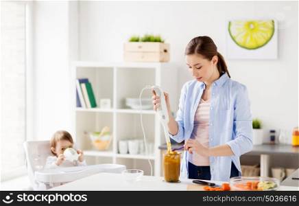 family, food, child, eating and parenthood concept - mother with blender cooking puree for her baby sitting in highchair at home kitchen. mother with blender cooking baby food at home