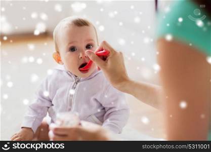 family, food, child, eating and parenthood concept - mother with puree and spoon feeding little baby at home over snow. mother with spoon feeding little baby at home