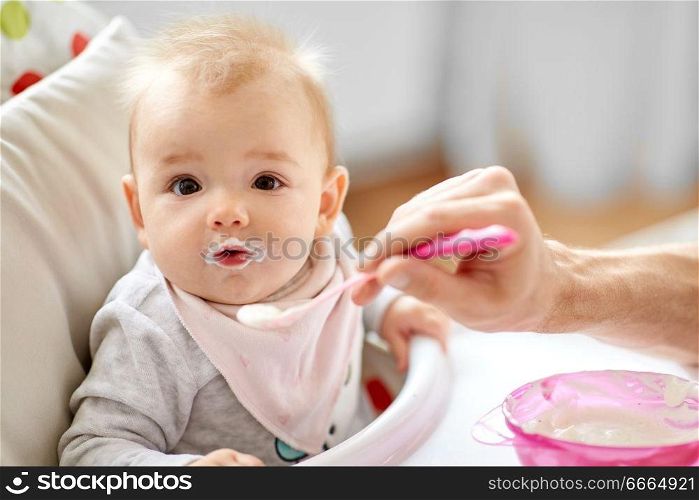 family, food, eating and people concept - father feeding little baby sitting in highchair with puree by spoon at home. father feeding baby sitting in highchair at home