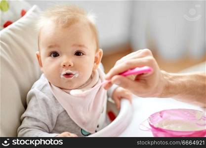 family, food, eating and people concept - father feeding little baby sitting in highchair with puree by spoon at home. father feeding baby sitting in highchair at home