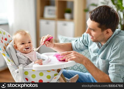 family, food, eating and people concept - happy father feeding little baby daughter sitting in highchair with puree by spoon at home. father feeding happy baby in highchair at home