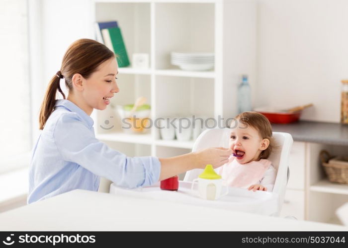 family, food, eating and people concept - happy mother with puree and spoon feeding little baby sitting in highchair at home. happy mother feeding baby with puree at home