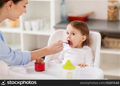 family, food, eating and people concept - happy mother with puree and spoon feeding little baby sitting in highchair at home. happy mother feeding baby with puree at home