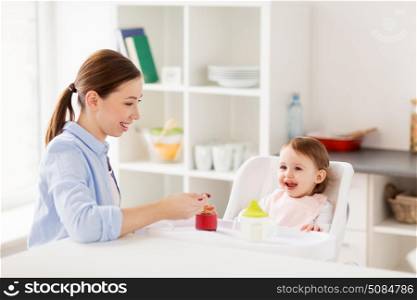 family, food, eating and people concept - happy mother with puree and spoon feeding little baby sitting in highchair at home. happy mother feeding baby with puree at home. happy mother feeding baby with puree at home