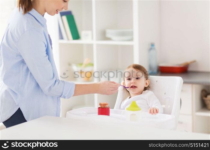 family, food, eating and people concept - happy mother with puree and spoon feeding little baby sitting in highchair at home. happy mother feeding baby with puree at home