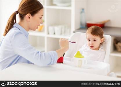 family, food, eating and people concept - happy mother with puree on spoon feeding little baby sitting in highchair at home. happy mother feeding baby with puree at home