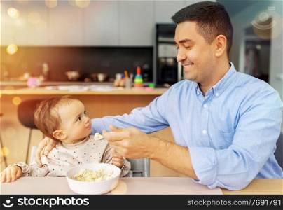 family, food, eating and people concept - happy smiling middle-aged father feeding little baby daughter sitting in highchair with puree by spoon at home. happy father feeding baby in highchair at home