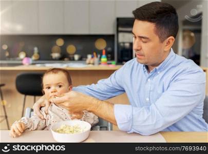 family, food, eating and people concept - middle-aged father feeding little baby daughter sitting in highchair with puree by spoon at home. middle-aged father feeding baby daughter at home