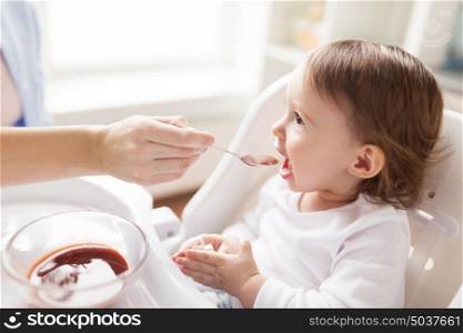 family, food, eating and people concept - mother with puree and spoon feeding little baby sitting in highchair at home. mother feeding baby with puree at home