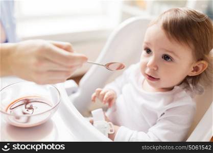family, food, eating and people concept - mother with puree and spoon feeding little baby sitting in highchair at home. mother feeding baby with puree at home