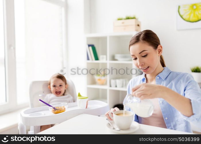 family, food, eating, breakfast and people concept - happy mother pouring milk from jug to coffee cup and little baby sitting in highchair at home kitchen. happy mother and baby having breakfast at home