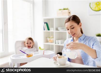 family, food, eating, breakfast and people concept - happy mother pouring milk from jug to coffee cup and little baby sitting in highchair at home kitchen. happy mother and baby having breakfast at home