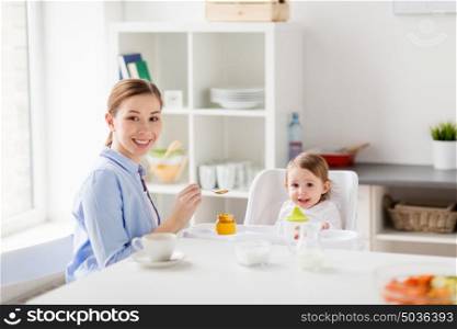 family, food, eating, breakfast and people concept - happy mother with puree and spoon feeding little baby sitting in highchair at home. happy mother feeding baby with puree at home