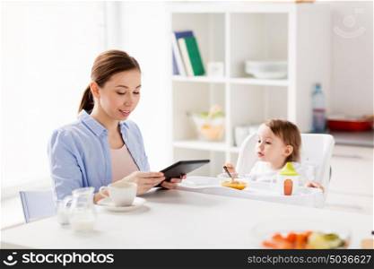 family, food, eating, breakfast and people concept - happy mother with tablet pc computer and little baby sitting in highchair at home kitchen. happy mother and baby having breakfast at home
