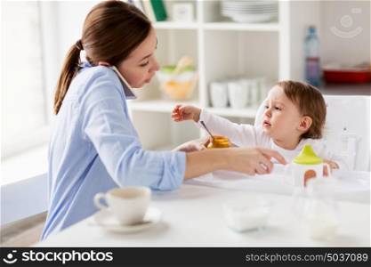family, food, eating, multitasking and people concept - mother with puree and spoon feeding little baby sitting in highchair and calling on smartphone at home. mother with smartphone feeding baby at home