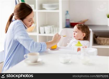 family, food, eating, multitasking and people concept - mother with puree and spoon feeding little baby sitting in highchair and calling on smartphone at home. mother with smartphone feeding baby at home