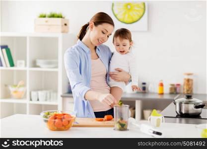 family, food, healthy eating, cooking and people concept - happy mother adding chopped vegetables to blender cup and holding little baby girl at home kitchen. happy mother and baby cooking food at home kitchen