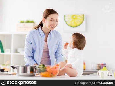 family, food, healthy eating, cooking and people concept - happy mother and little baby girl with carrot at home kitchen. happy mother and baby eating at home kitchen