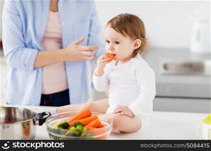 family, food, healthy eating, cooking and people concept - happy mother and little baby girl with carrot at home kitchen. happy mother and baby eating at home kitchen. happy mother and baby eating at home kitchen