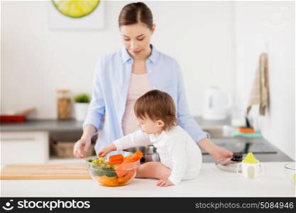 family, food, healthy eating, cooking and people concept - happy mother and little baby girl with bowl of vegetables at home kitchen. happy mother and baby with food at home kitchen. happy mother and baby with food at home kitchen