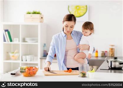 family, food, healthy eating, cooking and people concept - happy mother chopping carrot and little baby girl with vegetables and pot at home kitchen. happy mother and baby cooking food at home kitchen