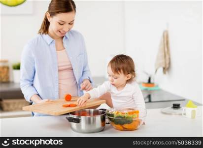 family, food, healthy eating, cooking and people concept - happy mother with chopped carrot and little baby girl with vegetables and pot at home kitchen. happy mother and baby cooking food at home kitchen