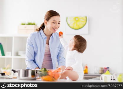 family, food, healthy eating, cooking and people concept - little baby feeding his mother with carrot at home kitchen. baby feeding mother with carrot at home kitchen