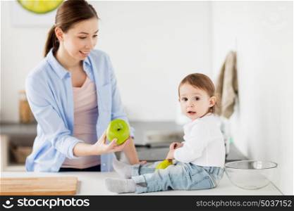 family, food, healthy eating, people and motherhood concept - happy young mother giving green apple to baby at home kitchen. mother giving green apple to baby at home kitchen