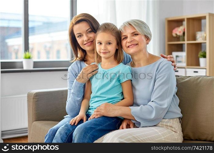 family, generation and female concept - portrait of smiling mother, daughter and grandmother sitting on sofa at home. portrait of mother, daughter and grandmother
