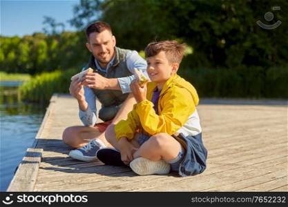 family, generation and food concept - happy smiling father and son with tablet pc computer eating sandwiches on river berth. father and son eating sandwiches on river berth