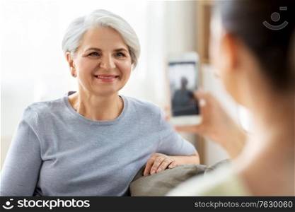 family, generation and people concept - adult daughter with smartphone photographing happy smiling senior mother at home. adult daughter photographing senior mother at home