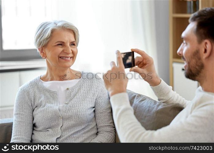 family, generation and people concept - adult son photographing smiling senior mother by smartphone at home. adult son photographing senior mother at home