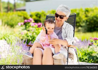 family, generation and people concept - happy grandmother with baby granddaughter calling on smartphone at summer garden. grandmother and baby granddaughter with smartphone