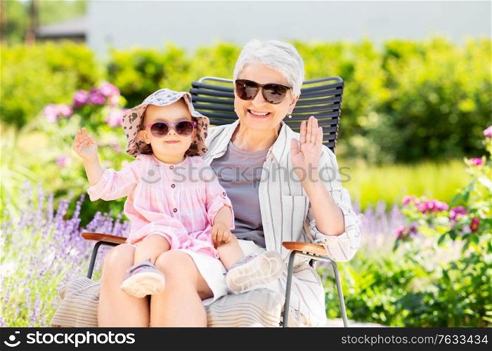 family, generation and people concept - happy grandmother with baby granddaughter waving hands at summer garden. happy grandmother and baby granddaughter at garden