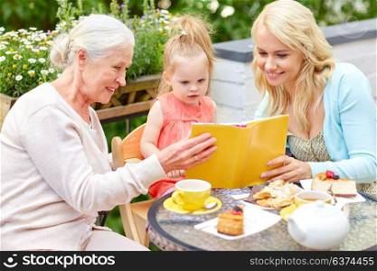 family, generation and people concept - happy mother with daughter and grandmother reading book at cafe or restaurant terrace. happy family reading book at cafe