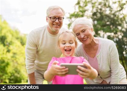 family, generation and people concept - happy smiling grandmother, grandfather and little granddaughter taking selfie by smartphone at park. senior grandparents and granddaughter selfie