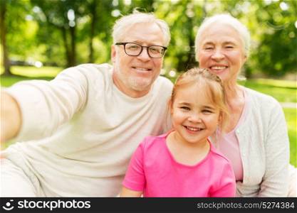 family, generation and people concept - happy smiling grandmother, grandfather and little granddaughter taking selfie at park. senior grandparents and granddaughter selfie