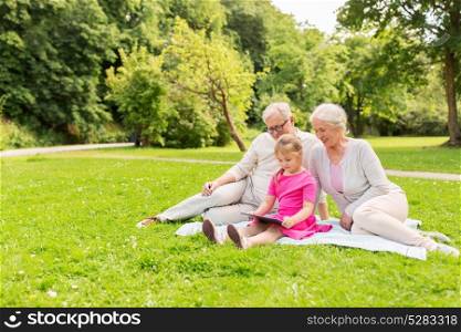 family, generation and people concept - happy smiling grandmother, grandfather and little granddaughter with tablet pc computer sitting on blanket at park. grandparents and granddaughter with tablet pc