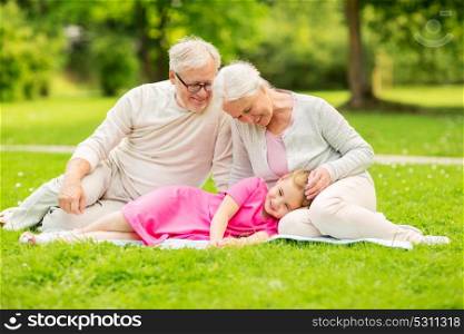 family, generation and people concept - happy smiling grandmother, grandfather and little granddaughter resting on blanket at park. senior grandparents and granddaughter at park