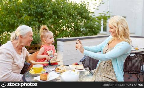 family, generation and people concept - happy smiling mother with smartphone photographing daughter and grandmother at cafe or restaurant terrace. woman photographing her family at cafe