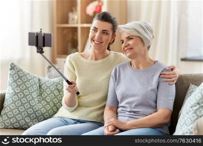 family, generation and people concept - happy smiling senior mother and adult daughter taking picture with smartphone on selfie stick at home. senior mother with daughter taking selfie at home