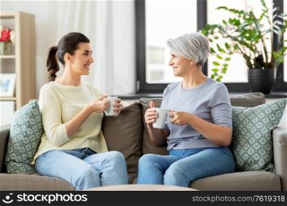 family, generation and people concept - happy smiling senior mother with adult daughter drinking coffee or tea and talking at home. senior mother and adult daughter drinking coffee
