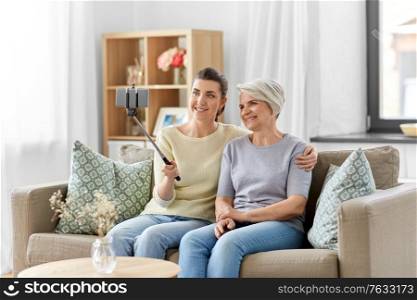 family, generation and people concept - happy smiling senior mother with adult daughter taking picture with smartphone on selfie stick at home. senior mother with daughter taking selfie at home