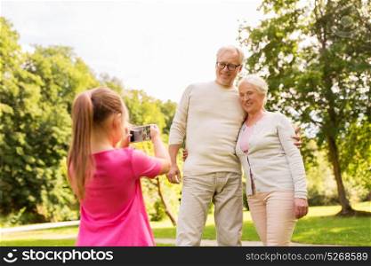 family, generation and people concept - little granddaughter photographing happy smiling grandmother and grandfather by smartphone at park. granddaughter photographing grandparents at park