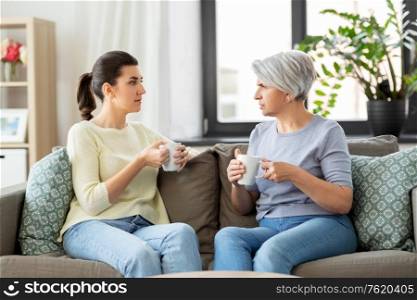 family, generation and people concept - senior mother with adult daughter drinking coffee or tea and talking at home. senior mother and adult daughter drinking coffee