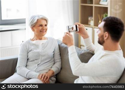 family, generation and people concept - smiling adult son photographing senior mother by smartphone at home. adult son photographing senior mother at home